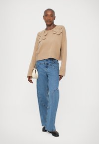 Beige stickad kort tröja med tre blommor som detaljer på axlarna, ihop med ljusblå vida jeans och en liten vit handväska.