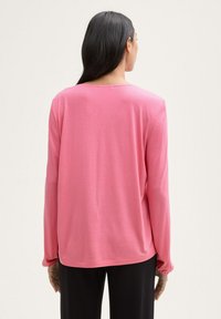 Top de manga larga rosa con cuello redondeado, textura suave y corte ligeramente relajado. Las mangas se recogen en las muñecas. Combinado con pantalones negros.