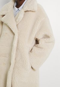 Abrigo de peluche beige con una superficie suave y texturizada, cuello con muesca, grandes botones y bolsillos laterales, que presenta un diseño de ajuste relajado.