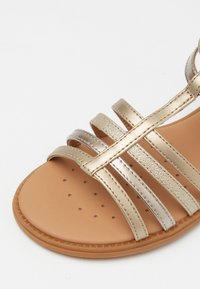 Arany fémes szandálok, több vékony pánttal és lapos, barna gumi talppal, amely perforációkkal készült a szellőzés érdekében.