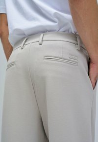 Pantalones de color beige claro con una tela texturizada, que cuentan con dos bolsillos traseros, una cintura con trabillas para el cinturón y una pequeña etiqueta de la marca.