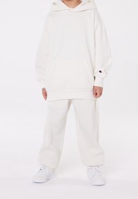 Barn i en överdimensionerad vit hoodie och matchande sweatpants med vita sneakers, som står mot en enkel vit bakgrund.