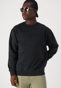 Μαύρη φούτερ μπλούζα με στρογγυλή λαιμόκοψη και ριμπ μανίκια, που διαθέτει μία υφασμάτινη λογότυπο Levi's στο στήθος, συνδυασμένη με ελιά-πράσινα παντελόνια και γυαλιά ηλίου.