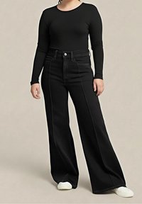 Femme portant un haut noir moulant à manches longues avec un pantalon noir taille haute à jambes larges et des baskets blanches, debout devant un fond uni.