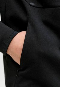 Rankos artinimas juodoje drabužio medžiagoje su tekstūruota audinio tekstūra, šonine kišene su užtrauktuku ir ribbingo manžetės detale.