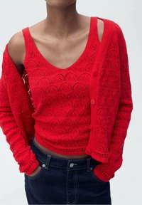 Rode gebreide cardigan met een v-hals, decoratief opengewerkt patroon en knoopsluiting. Draagt over een bijpassend rood mouwloos topje.