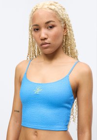 Νεαρή γυναίκα με μακριά ξανθά κοτσίδια, που φοράει μια υφασμάτινη μπλε κοντή μπλούζα με λεπτές τιράντες και ένα μικρό κεντητό λογότυπο.