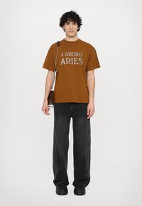 Brown cotton t-shirt with "J'ADORE ARIES" graphic, paired with black wide-leg jeans featuring side stitching. Black footwear completes the look.