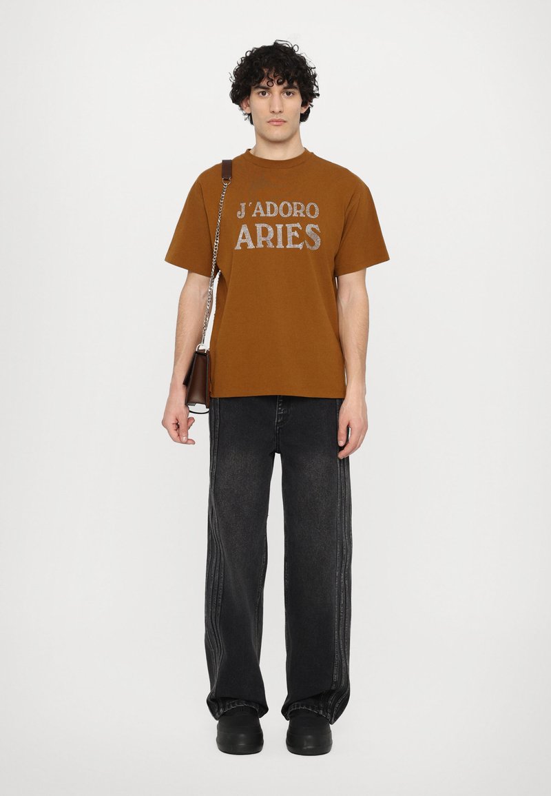 Brown cotton t-shirt with "J'ADORE ARIES" graphic, paired with black wide-leg jeans featuring side stitching. Black footwear completes the look.