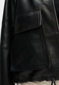 Κοντινό πλάνο τσέπης μαύρου δερμάτινου μπουφάν με καπάκι και εμφανείς ραφές, δίπλα σε ασημένιο φερμουάρ και στερεωμένο στρίφωμα με κορδόνι.