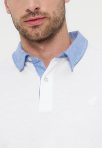 Άνδρας με κοντά γένια που φοράει λευκό πόλο μπλουζάκι με ανοιχτογάλανο γιακά και δύο κουμπιά στο άνοιγμα, που δείχνει το άνω μέρος του στήθους και του λαιμού.