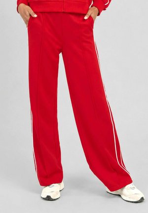 Personne portant un pantalon de survêtement rouge ample avec des bandes latérales blanches et des baskets blanches, les mains dans les poches, debout sur un sol gris.