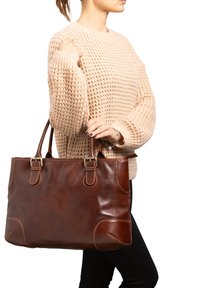 Borsa tote in pelle marrone con manici doppi, caratterizzata da una superficie liscia e cuciture a contrasto. Indossata con un maglione lavorato a maglia beige spesso.