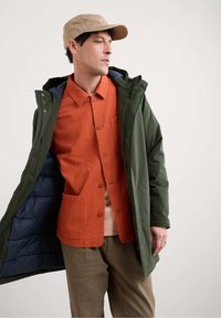 Zelený nepremokavý parka s kapucňou, cez oranžovú košeľu na gombíky s náprsníkovou vreckom, nosená s béžovou čiapkou a textúrovanými hnedými nohavicami.