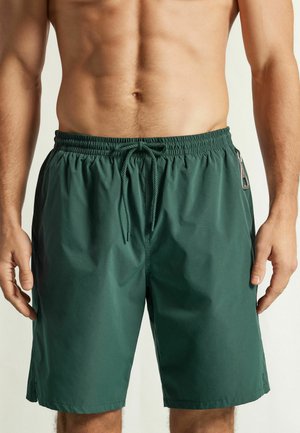 Torse masculin portant un short de bain vert foncé avec taille à cordon et poche latérale sur un fond uni.
