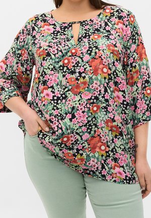 Blomstret bluse med sort baggrund, prydet med lyserøde og røde blomster, grønne blade og en lille nøglehul-udskæring. Matchet med lysegrønne bukser.