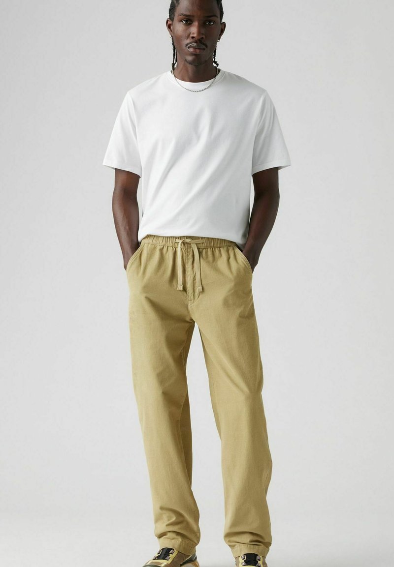 Camiseta blanca de manga corta combinada con pantalones elásticos de color caqui claro. El modelo está de pie con las manos en los bolsillos, llevando zapatos de diseño.