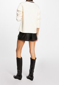 Donna che indossa un maglione bianco a maniche lunghe, pantaloni corti in pelle nera e stivali neri al ginocchio, in piedi con la schiena rivolta verso la fotocamera.