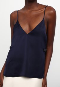 Camisole en soie bleu marine avec des bretelles fines, décolleté en V et accents sur les côtés ornés de petits éléments en métal noir. Texture lisse et design fluide.