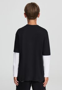 Czarny t-shirt z krótkim rękawem noszony na długim białym rękawie, z okrągłym dekoltem i luźnym krojem, widziany z tyłu.