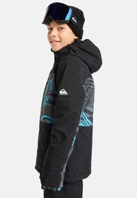 Schwarze Skijacke mit blauen Akzenten und grafischen Mustern, mit Kapuze und Quiksilver-Logo, getragen mit einer Mütze und einer Skibrille.