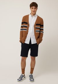 Brun stickad cardigan med navy, vitt och orange randning, bärs över en vit skjorta med krage. Kombineras med mörka shorts och sneakers.