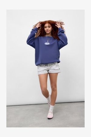 Purple oversized sweatshirt with a front logo, paired with light gray shorts. Model wears white socks and light pink shoes. Neutral background.