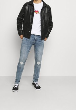 Muž v černé kožené bundě, bílém tričku s motivem srdce, roztrhaných modrých úzkých džínách a černobílých teniskách stojí opřený o bílou zeď.