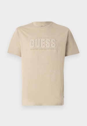 Beige cotton t-shirt with a crew neck, featuring embossed "GUESS" logo and "CLASSIC AMERICAN STYLE" text in lighter color. Short sleeves.