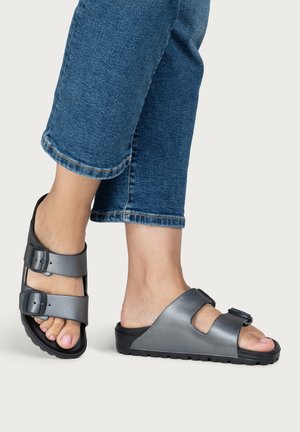 Feet wearing metallic grey double-strap slide sandals paired with cropped blue denim jeans on a light background.