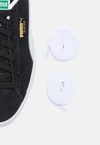 Black suede sneaker with white rubber sole and black laces, accompanied by two coiled white shoelaces. Gold brand logo on side.