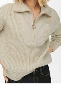 Pull en tricot beige avec un col haut, doté d'une fermeture éclair quart de tour et d'un motif texturé, confectionné dans un matériau doux.