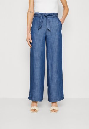 Niebieskie spodnie z wysokim stanem i szerokimi nogawkami z materiałowym paskiem, noszone z białymi plecionymi sandałami i białym bezrękawnikiem.