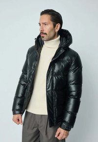 Veste noire en duvet avec capuche, présentant une texture brillante, fermeture éclair à l'avant et poches latérales. Stylée avec un col roulé crème clair en dessous.