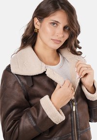 Chaqueta de shearling marrón con un collar y puños suaves en beige. Presenta una cremallera dorada y detalles de cuero en las mangas. Diseño clásico de bomber.