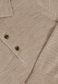 Beige kötött pólóing, texturált felülettel. Két sötét gomb található, arany díszítéssel a gallér közelében. Klasszikus dizájn és testhezálló fazon.