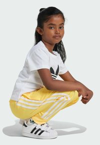 Maglietta bianca con logo nero, pantaloni tuta gialli con stripe bianche e scarpe da ginnastica in bianco e nero. I capelli sono lunghi e scuri.