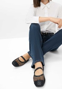 Femme assise en tailleur portant un jean bleu foncé, une chemise blanche boutonnée et des ballerines Mary Jane noires en maille avec des brides élastiques.