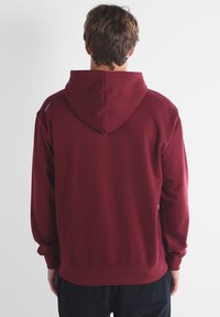 Hoodie bordeaux avec capuche à cordon de serrage, poignets et ourlet côtelés, fabriqué en tissu doux. Marque minimaliste appliquée près de l'épaule.