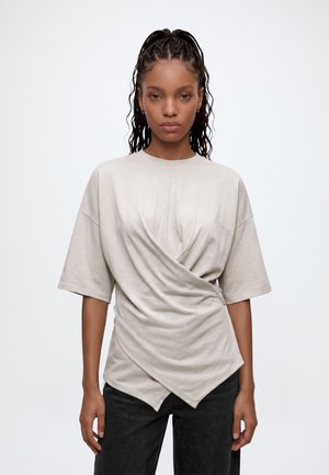 Femme aux longs cheveux tressés portant un haut beige style cache-cœur à manches courtes et un pantalon noir, debout devant un fond blanc uni.