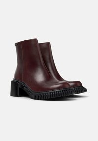 Botas de tobillo de cuero burdeos con una suela gruesa de goma negra, que presentan una puntera redondeada y textura lisa. Diseño minimalista sin cierres visibles.