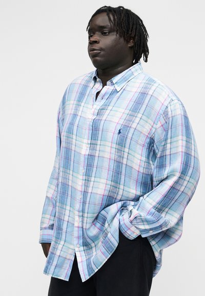 Persona de piel oscura y rastas que lleva una camisa holgada de manga larga a cuadros en tonos pastel, con la mano en el bolsillo, de pie frente a un fondo blanco.