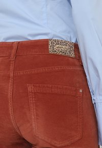 Personne portant un pantalon couleur rouille avec une poche arrière et une étiquette de marque à imprimé léopard, associée à une chemise bleu clair.