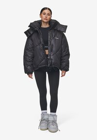 Μαύρο μπουφάν πούφφερ με ψηλό γιακά, λαμπερή υφή και oversized εφαρμογή. Φορεμένο πάνω από μια μαύρη crop top μπλούζα και συνδυασμένο με μαύρα κολάν και γκρι μπότες.