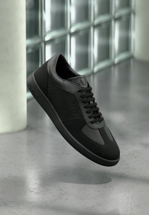 Czarny niski sneaker z teksturowanymi bocznymi panelami i okrągłym czubkiem, unoszący się nad polerowaną posadzką betonową w pobliżu ściany z bloków szklanych.