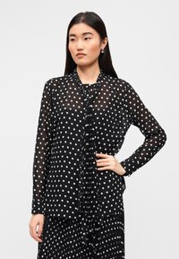 Μαύρη διαφανής μπλούζα με λευκές πουά, μακριά μανίκια και πιετές κορδόνι στο ντεκολτέ. Υφή υφάσματος με απαλό πέσιμο.