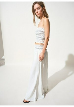 Mlada žena stoji u bijelom topu bez naramenica s crnim šavovima i širokim bijelim hlačama, okrenuta u profil na jednobojnoj pozadini.