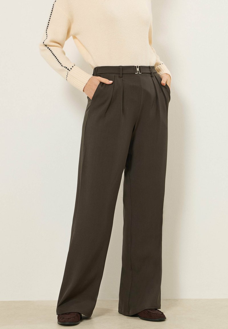 Vrouw draagt een donkerbruine wijde broek met hoge taille en riem, een crèmekleurige trui met zwarte stikseldetails, handen in de zakken, bruine schoenen zichtbaar.