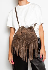 Borsa a secchiello in suede marrone con dettagli in frange, chiusura con coulisse e tracolla regolabile, indossata con una maglietta bianca e pantaloni neri.
