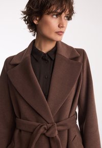 Brązowy płaszcz z paskiem, z klapami szalowymi, teksturowana tkanina i dopasowany krój. Noszony na ciemnej koszuli z guzikami i kołnierzykiem.